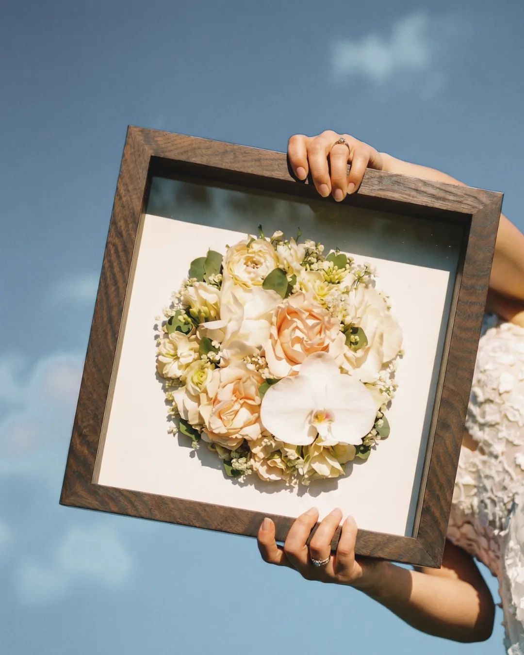 Bouquet de mariée préservé dans un cadre en bois tenu contre le ciel, campagne L'écrin pour Les Joyaux Purs