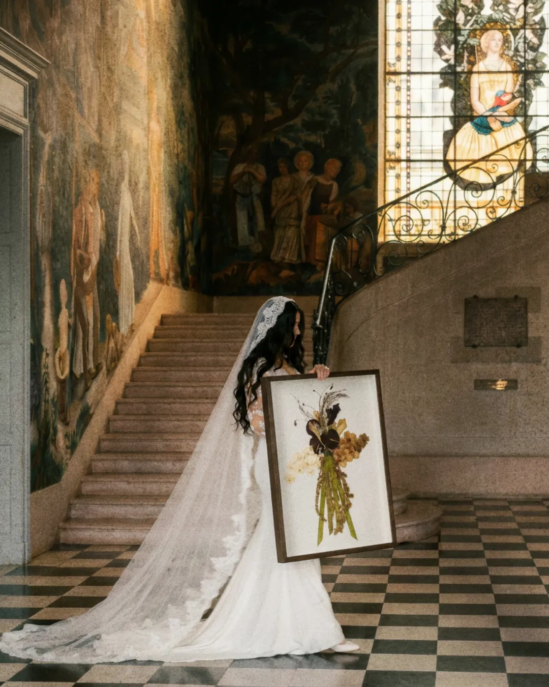 Mariée portant son tableau floral au pied d'un escalier sous un vitrail